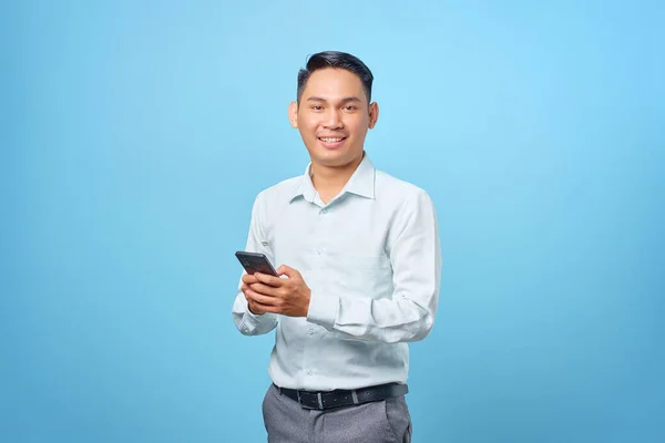 Gülümseyen yakışıklı iş adamı elinde akıllı telefonla mavi arka plandaki kameraya bakıyor.