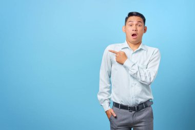 Şok olmuş genç yakışıklı işadamı mavi arka plandaki fotokopi alanını işaret ediyor