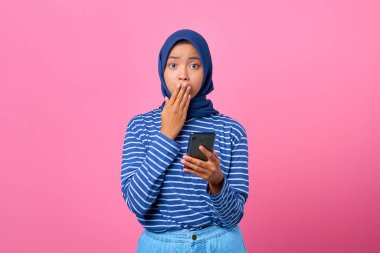 Şok olmuş Asyalı genç bir kadının portresi. Elinde akıllı telefon ve eliyle ağzını kapatıyor.