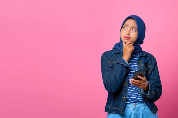 Portrait of young Asian woman thinking about question with hand on chin while holding smartphone