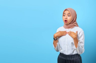 Şok olmuş genç Asyalı bir kadının portresi, ağzı açık ve elini göğsüne koyup mavi arka plan üzerine koydu.