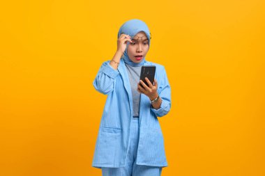 Sarı arka planda izole edilmiş cep telefonu ve gözlük kullanan genç Asyalı kadın şok oldu.