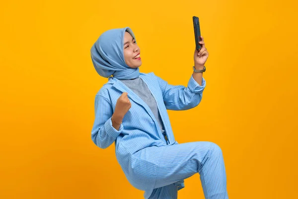 Neşeli genç Asyalı kadın cep telefonu tutuyor ve sarı arka planda başarıyı kutluyor.