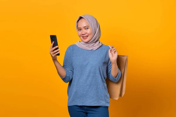 Sarı Arka Planda Telefonu Alışveriş Çantasını Tutan Neşeli Asyalı Kadın — Stok fotoğraf