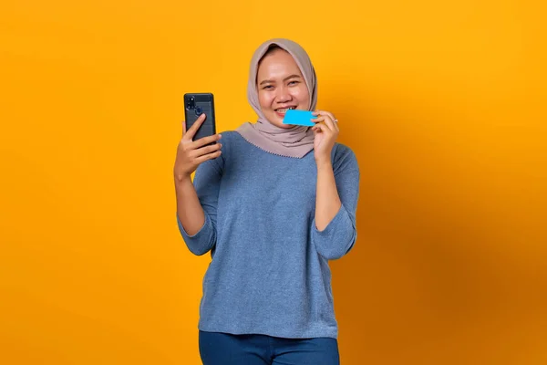 Heyecanlı Asyalı kadın akıllı telefon tutuyor ve sarı arka planda kredi kartını ısırıyor.