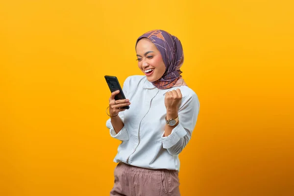 Heyecanlı Asyalı kadın cep telefonuyla sarı arka planda şansı kutluyor.