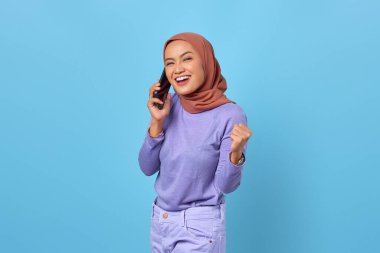 Neşeli genç Asyalı kadın mavi arka planda zaferi kutlarken cep telefonuyla konuşuyor.
