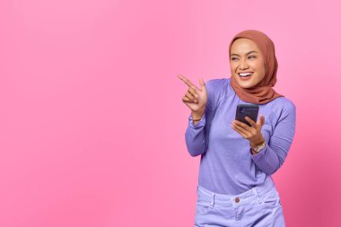 Elinde cep telefonu olan gülümseyen Asyalı kadın pembe arka plandaki fotokopi alanını işaret ediyor.