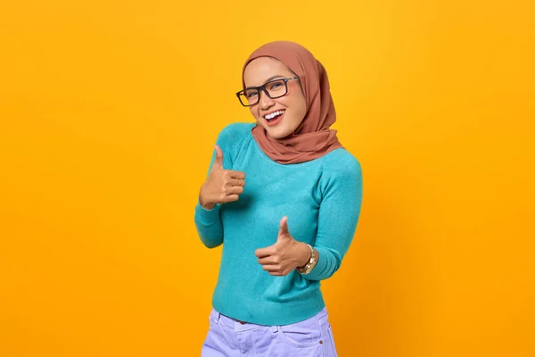 Neşeli, güzel, genç Asyalı kadın sarı arka planda izole edilmiş baş parmak hareketi gösteriyor.