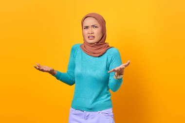 Kafası karışmış genç Asyalı kadın omuz silkiyor sarı arka planda el kaldırıyor.