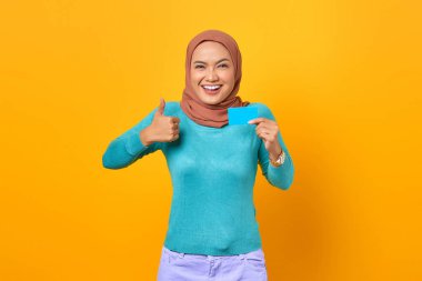 Gülümseyen genç Asyalı kadın kredi kartı tutuyor ve sarı arka planda baş parmak işareti yapıyor.