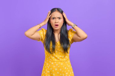 Şaşırmış, ağzı açık, kafası mor arka planda iki eliyle tutulan Asyalı bir kadın.