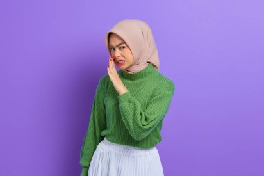 Güzel Asyalı genç bir kadın, yeşil süveter giymiş, eli ağızda, sırlarını anlatıyor. Mor arka planda izole edilmiş, fısıldayan bir dedikodu.