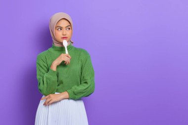 Aç, yeşil kazaklı, elinde kaşıkla dolaşan güzel bir Asyalı kadın mor arka planda izole edilmiş lezzetli bir yemek hayal ediyor.