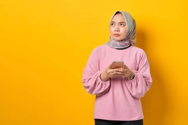 Pembe tişörtlü, akıllı telefon kullanan düşünceli genç Asyalı kadın sarı arka planda bir soru düşünüyor.