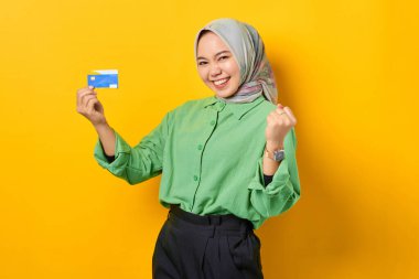 Yeşil tişörtlü, elinde kredi kartı olan heyecanlı genç Asyalı kadın sarı arka planda birinci sınıf jest yapıyor.