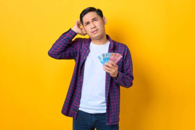 Ekose gömlekli kafası karışmış genç Asyalı adam sarı arka planda izole edilmiş para banknotları tutuyor.