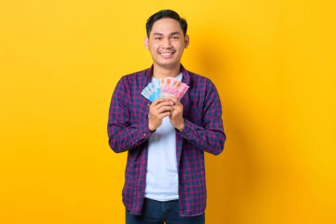 Gülümseyen, ekose gömlekli genç Asyalı adam sarı arka planda izole edilmiş para banknotları tutuyor.