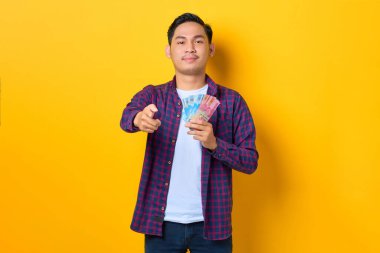 Ekose gömlekli gülümseyen genç Asyalı adam elinde para banknotları ve sarı arka planda izole edilmiş kamerayı işaret ediyor.