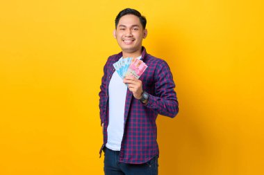 Gülümseyen, ekose gömlekli genç Asyalı adam sarı arka planda izole edilmiş para banknotları tutuyor.