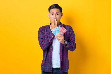 Ekose gömlekli, elinde para banknotları ve sarı arka planda elleri izole edilmiş ağzı olan Asyalı genç şoka uğramış bir adam.