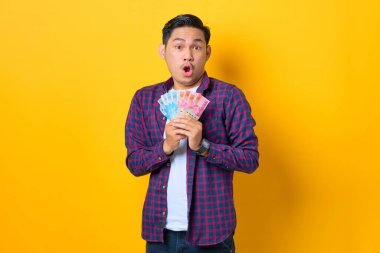 Ekose gömlekli, ağzı açık, sarı arka planda izole edilmiş para banknotları tutan Asyalı bir adam.