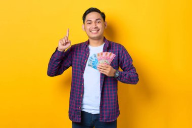 Ekose gömlekli neşeli genç Asyalı adam elinde para banknotları ve sarı arka planda izole edilmiş parmaklar tutuyor.