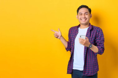 Ekose gömlekli neşeli genç Asyalı adam cep telefonu tutuyor ve sarı arka planda izole edilmiş fotokopi alanını işaret ediyor.