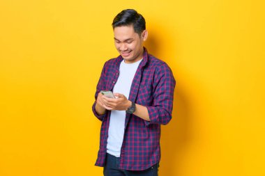 Ekose gömlekli gülümseyen genç Asyalı cep telefonu kullanıyor sarı arkaplanda izole edilmiş SMS mesajı yazıyor.