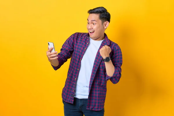 Ekose gömlekli heyecanlı genç Asyalı adam akıllı telefon kullanıyor ve sarı arka planda izole edilmiş kazanan hareketi yapıyor.