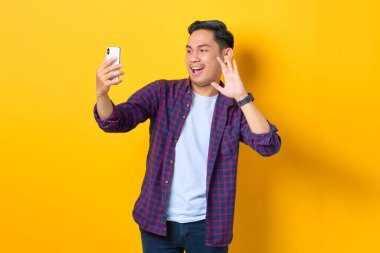 Ekose gömlekli neşeli genç Asyalı adam sarı arka planda el sallayarak cep telefonuyla selfie çekiyor.