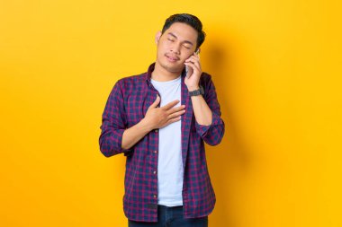 Ekose gömlekli memnun Asyalı genç adam sarı arka planda cep telefonuyla konuşuyor.
