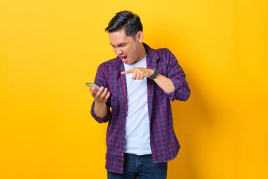 Ekose gömlekli kızgın genç Asyalı adam cep telefonu kullanıyor ve sarı arka planda izole bir şekilde bağırıyor.