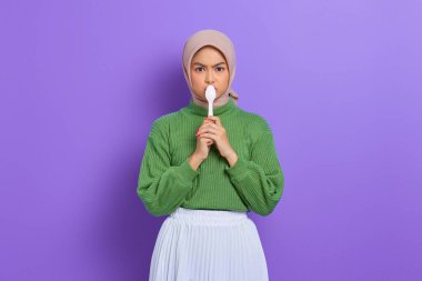 Yeşil kazaklı ve tesettürlü güzel Asyalı kadın, bir kaşıkla ağzını kapatıyor, mor arka planda izole edilmiş lezzetli yemekleri denemek istiyor.