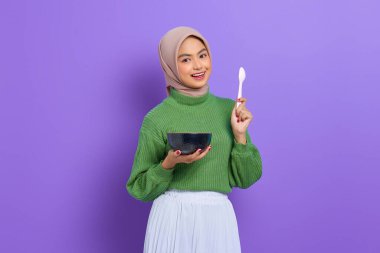 Gülümseyen, yeşil kazaklı, tesettürlü, tesettürlü, elinde kaşık ve kâseli, mor arka planda tahıl gevreği olan güzel Asyalı kadın.