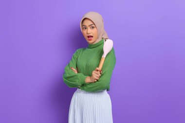 Şok olmuş yeşil kazaklı ve tesettürlü güzel Asyalı kadın mor arka planda izole edilmiş ev işlerini yaparken mutfak aletlerini tutuyor.