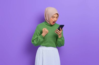Yeşil kazaklı ve tesettürlü sinirli genç Asyalı kadın mor arka planda izole bir şekilde yüksek sesle bağırırken akıllı telefon kullanıyor.