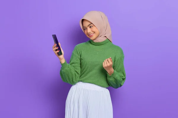 Heyecanlı yeşil kazaklı ve tesettürlü güzel Asyalı kadın cep telefonu kullanıyor, başarıyı kutluyor, mor arka planda iyi haberler alıyor.