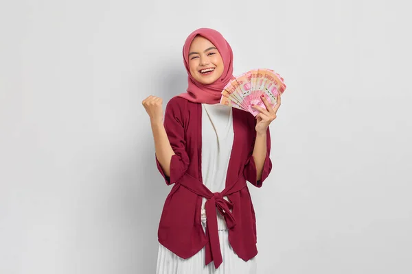 カジュアルなシャツとヒジャーブの陽気な美しいアジアの女性は 白い背景に隔離された幸運を祝う インドネシアルピア紙幣を示しています 人々の宗教生活の概念 — ストック写真