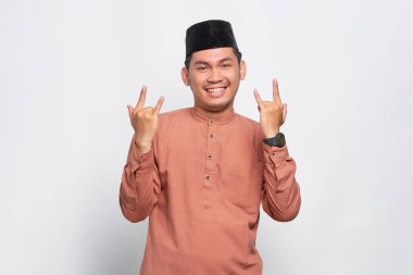 Heyecanlı Asyalı Müslüman adam bağırıyor ve elleri yukarıda beyaz arka planda rock sembolü yapıyor.