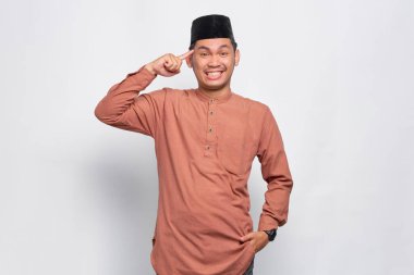 Gülümseyen Asyalı genç Müslüman adam, beyaz arka planda izole edilmiş parmaklarıyla başını işaret ediyor.