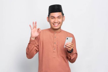 Gülümseyen Asyalı Müslüman bir adam cep telefonu tutuyor ve beyaz arka planda izole edilmiş bir işaret gösteriyor.