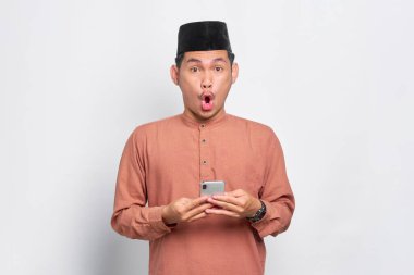 Asyalı genç Müslüman adam beyaz arka planda izole edilmiş ağzı açık bir cep telefonu kullanıyor.