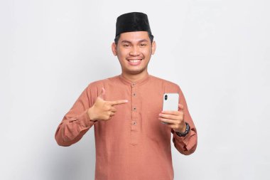 Gülümseyen Asyalı genç Müslüman adam beyaz arka planda izole edilmiş cep telefonunu işaret ediyor.