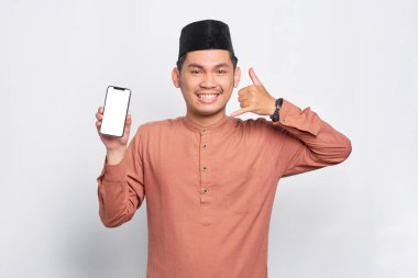 Gülümseyen Asyalı genç Müslüman adam cep telefonunu boş ekranda gösteriyor. Uygulama öneriyor ve işaret ediyor. Beyaz arka planda izole edilmiş bir şekilde beni arıyor.