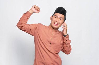 Heyecanlı Asyalı genç Müslüman adam cep telefonuyla konuşuyor ve kazanan jest yapıyor beyaz arka planda izole edilmiş.