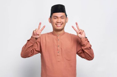 Gülümseyen Asyalı Müslüman adam beyaz arka planda izole edilmiş parmaklarıyla barış işareti gösteriyor