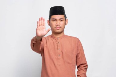 Asyalı, ciddi, genç Müslüman bir adam elini uzatmış, beyaz arka planda izole edilmiş dur hareketi gösteriyor.