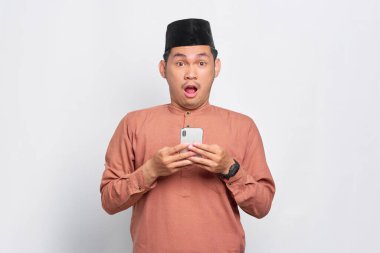 Asyalı genç Müslüman bir adam cep telefonu kullanıyor ve beyaz arka planda izole edilmiş kameraya bakıyor.