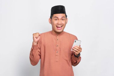 Heyecanlı Asyalı genç Müslüman bir adam cep telefonu kullanıyor ve başarıyı kutluyor, beyaz arka planda iyi haberler alıyor.